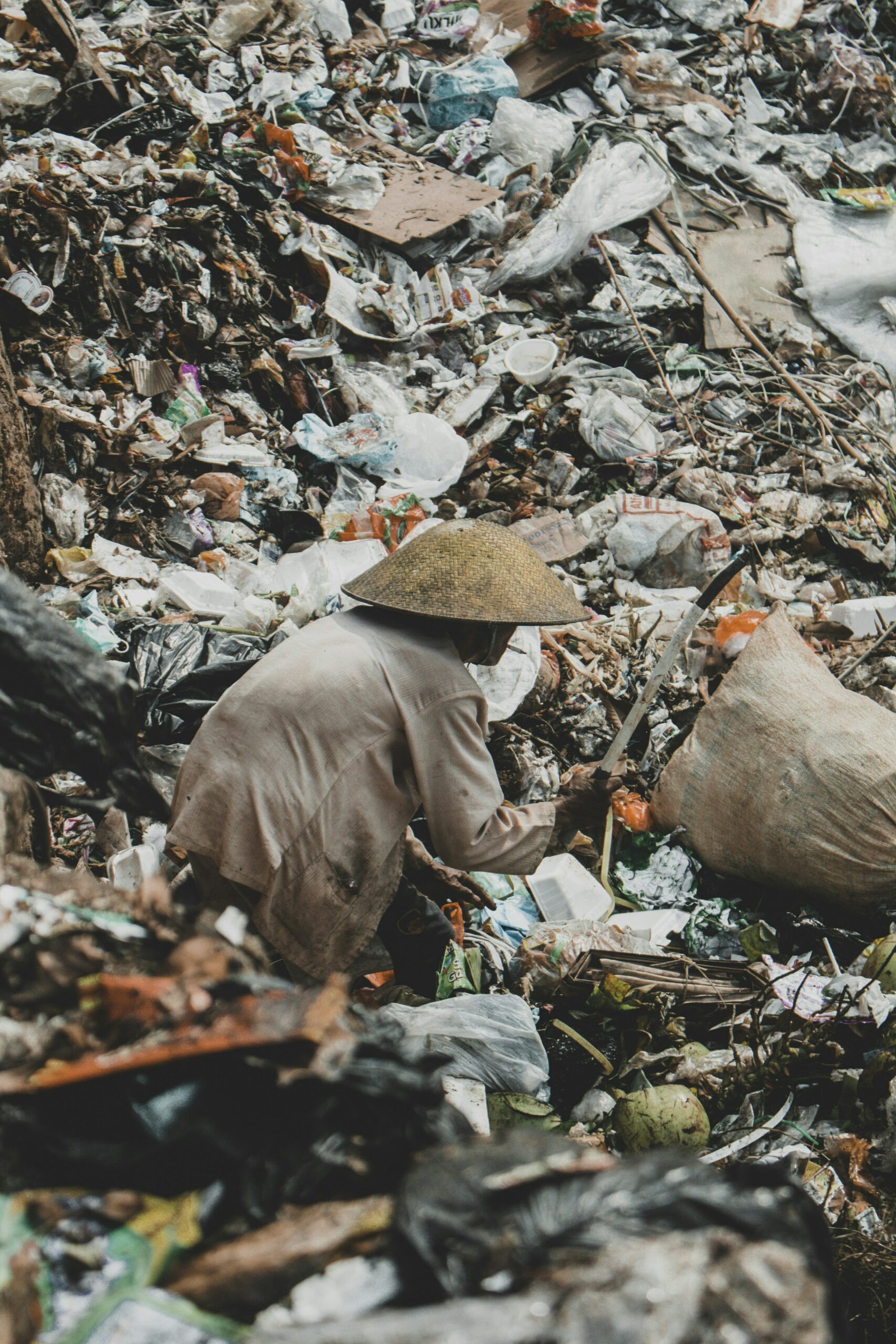 Photo of person in landfill. They are wearing a broad brim hat and appear to be working in some way to find valued items in the trash.